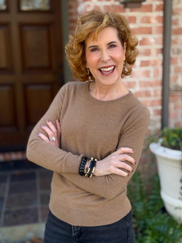 woman over 50 standing in front of a house wearing a mocha mousse cashmere sweater and black and gold bracelets