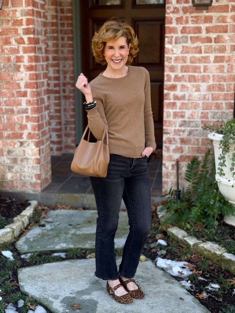 woman over 50 standing in front of a house wearing a mocha mousse cashmere sweater and black and gold bracelets and black jeans with leopard print mary jane shoes carrying a brown mocha mousse tote