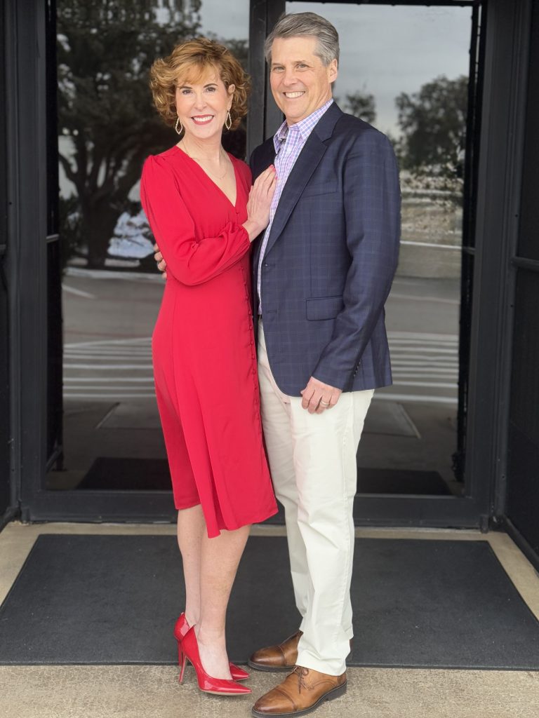 couple in an empty nester marriage posing in front of black doors at a restaurant. wife wearing red dress and man wearing blazer and slacks.