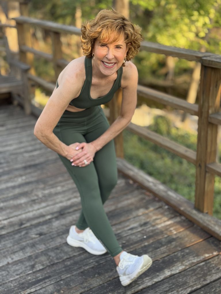 woman over 50 wearing definite articles sports bra and leggings standing on a wooden deck and stretching her right hamstring muscle