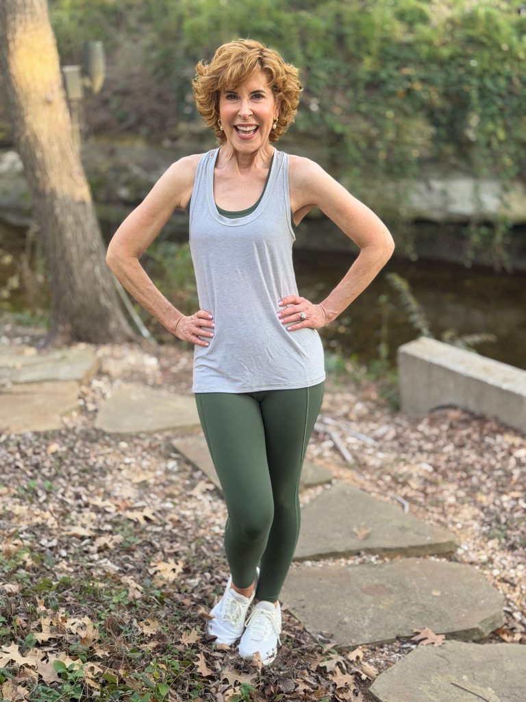 woman over 50 wearing definite articles sports bra, tank top, and leggings standing on a stone walkway by a stream