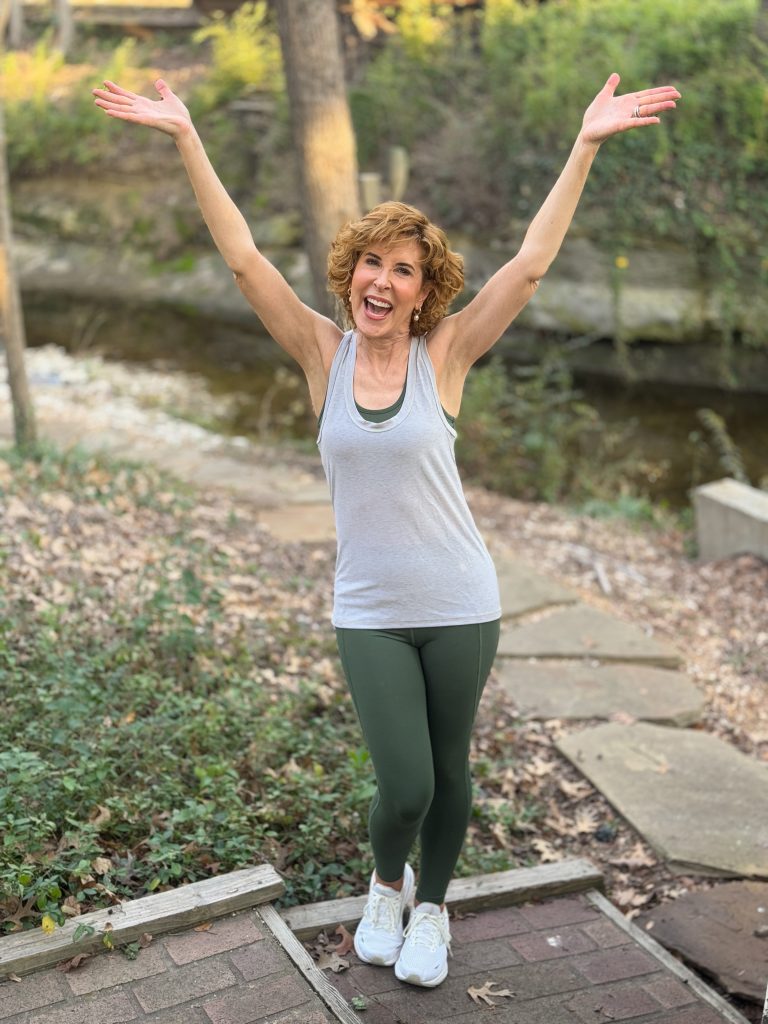 woman over 50 wearing definite articles sports bra, tank top, and leggings standing on a stone walkway by a stream with arms raised in the air