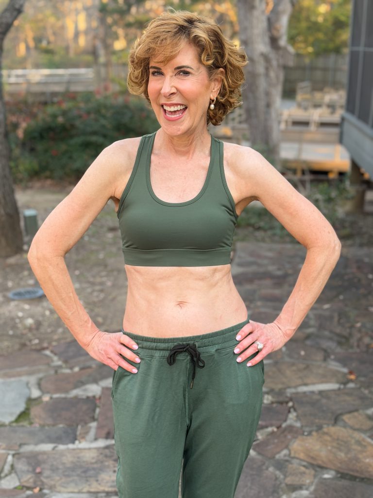 woman over 50 wearing definite articles sports bra and joggers standing on a stone walkway in a wooded area