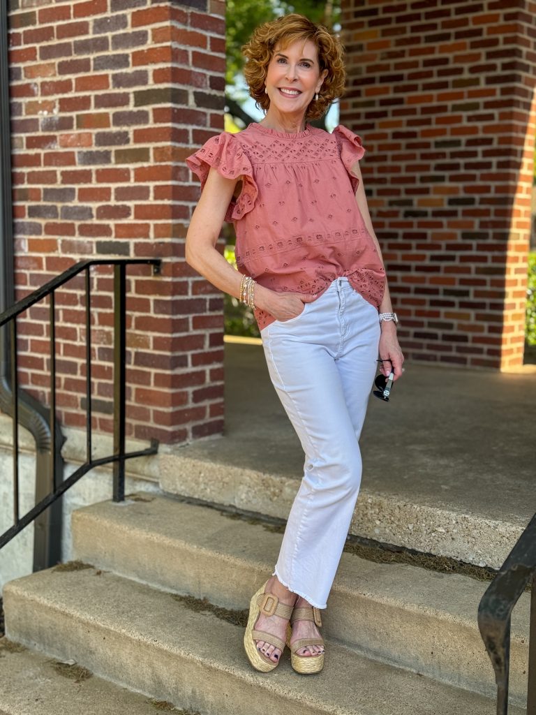 woman over 50 wearing coral eyelet top and white jeans standing outdoors on a staircase