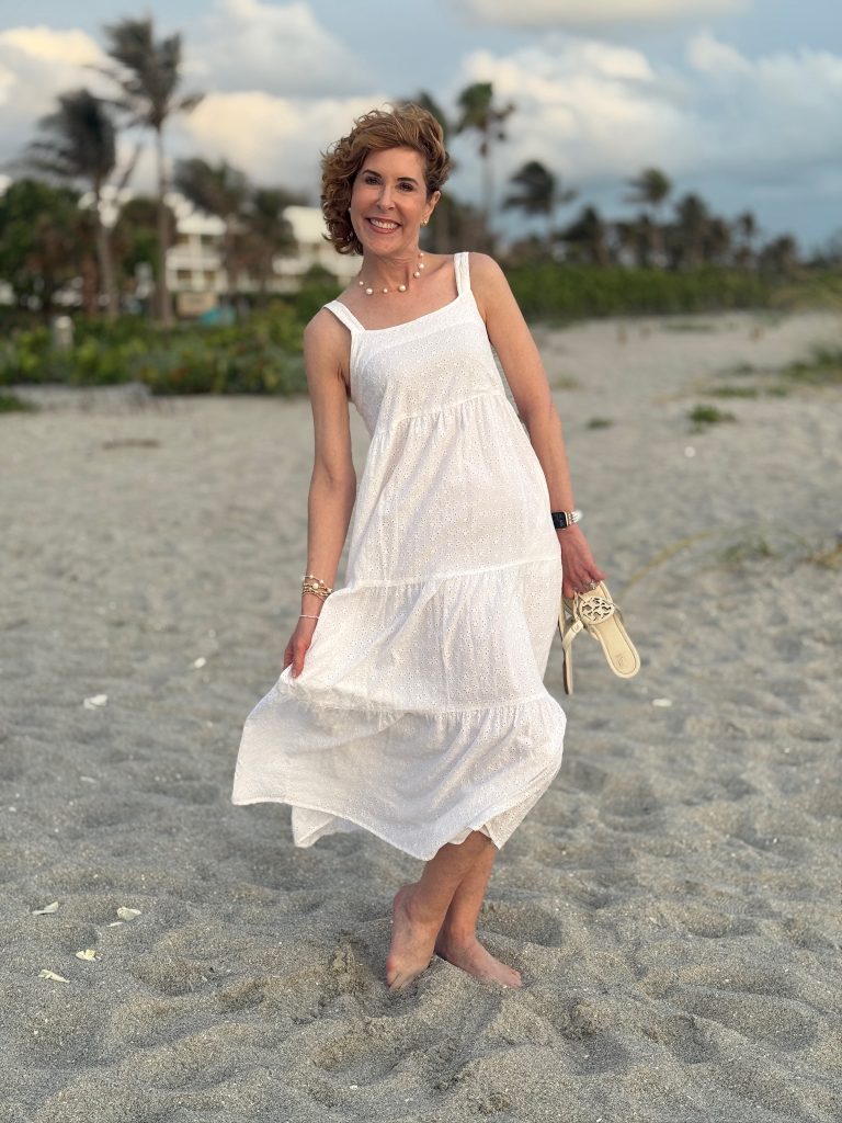 woman standing on a beach wearing a white eyelet sundress from amazon and holding a pair of sandals