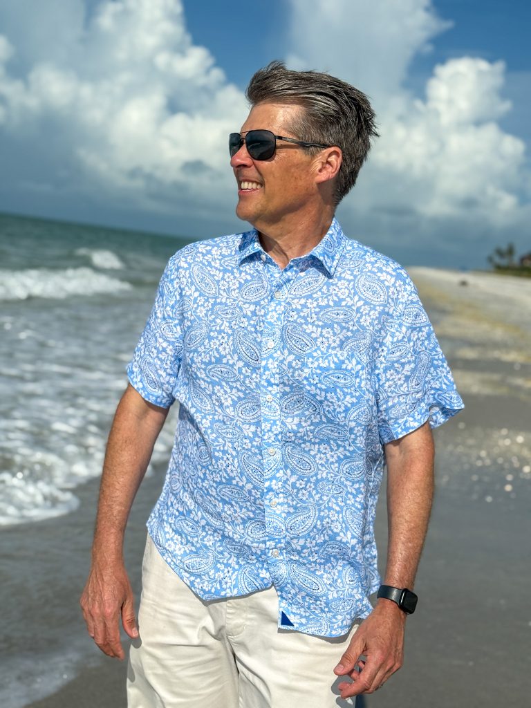 man standing on beach with blue and white linen shirt and white shorts