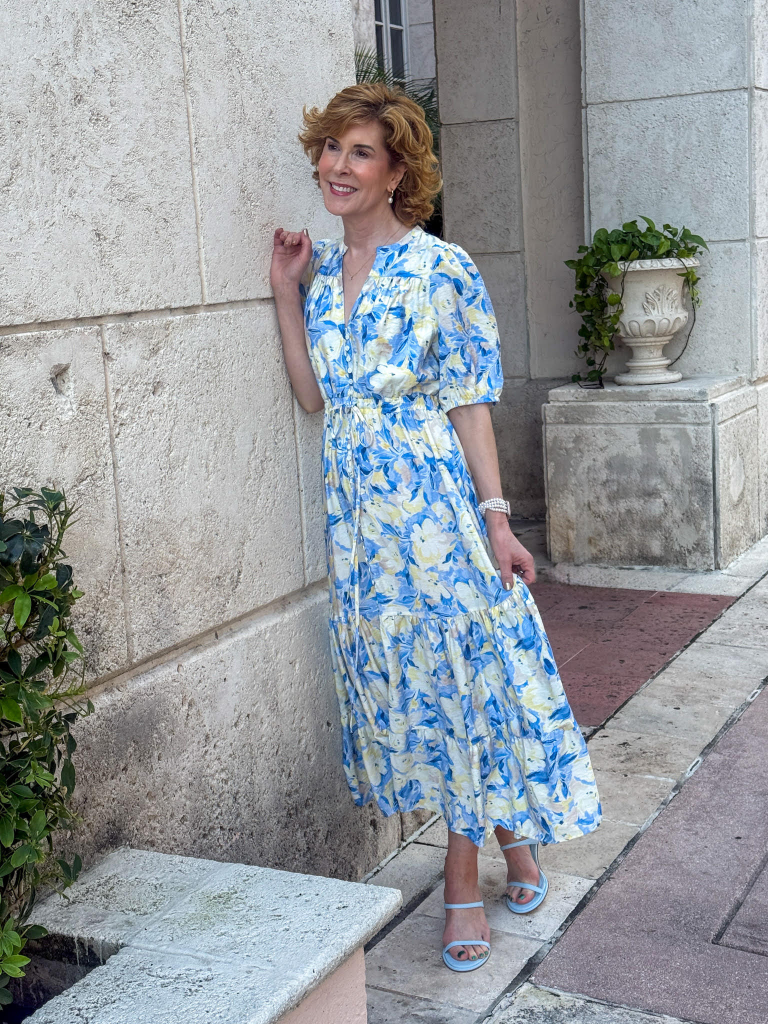 Woman standing beside a textured stone wall wearing a blue and pale yellow floral midi dress with elbow-length puff sleeves, a drawstring waist, and a tiered skirt. She pairs the dress with light blue strappy sandals and pearl bracelets, creating a soft, feminine spring outfit.