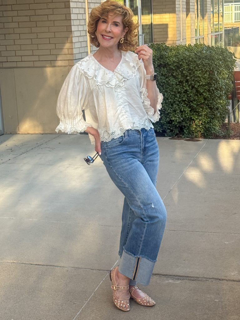 Woman wearing a cream lace blouse with puff sleeves and blue cuffed jeans, styled with tan studded flats, posing outdoors.