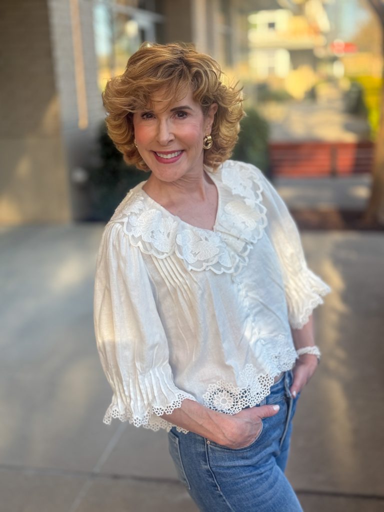 Woman wearing a cream lace blouse with a scalloped collar and puff sleeves, paired with high-waisted blue jeans, smiling outdoors.