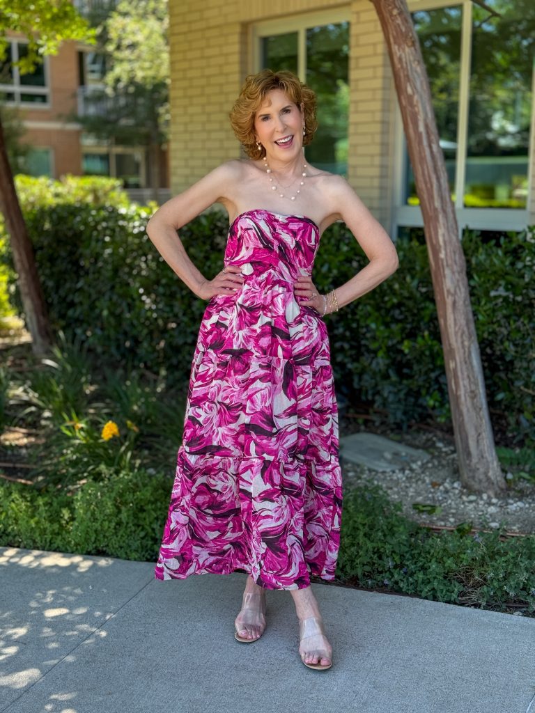 Smiling woman outdoors wearing a strapless pink floral midi dress with a full skirt, clear heeled sandals, and delicate jewelry, posing on a sunny sidewalk in front of greenery and a building.