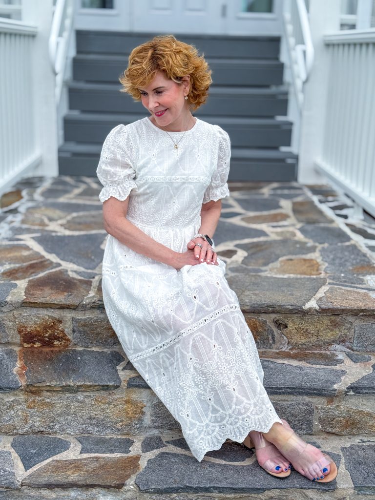 Woman wearing a white embroidered eyelet maxi dress sits on a stone wall in front of white steps, smiling as she looks down. She pairs the feminine summer dress with clear slide sandals and simple jewelry for a classic warm-weather outfit.
